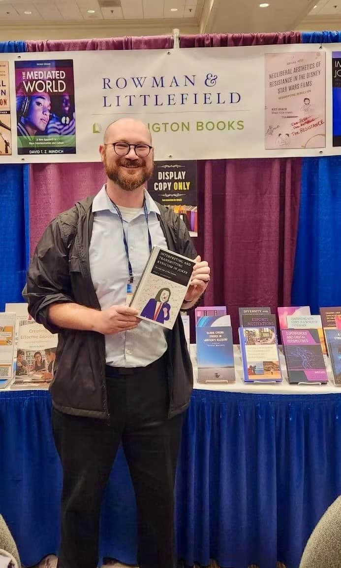 Dr. Kyle Hammonds holding a copy of Interpreting and Transmitting Kynicism in Joker at the Rowman & Littlefield booth at the National Communication Association convention